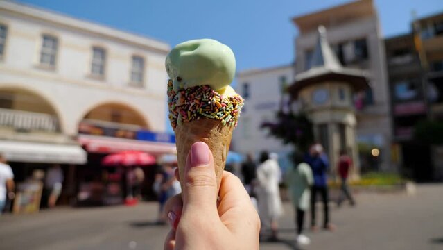 Tourist woman holds cone of ice cream, clip capturing tempting and flavorsome delight against blurred view of Buyukada clock square. This street food indulgence adds sweet touch to vibrant atmosphere
