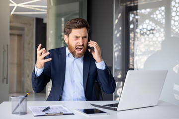 Angry young male businessman sitting at a desk in the office and talking aggressively on the phone,...