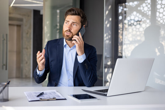 A serious young man businessman sits at an office table in a suit and discusses business on the phone with clients and partners