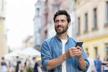 A smiling young man walks down the city street in casual clothes and holds a mobile phone in his...