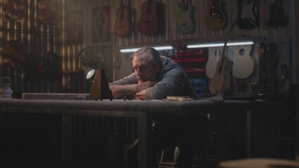 Fototapeta na wymiar Tired male artisan takes off glasses, looks at metronome. Mature carpenter relaxes after long workday in modern workshop for making musical instruments. Handcrafted wooden guitars hang on the wall.