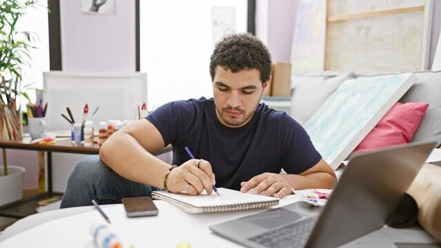 Young man with beard writing in notebook at home studio with laptop and art supplies.