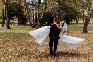 Stylish groom holds beautiful bride in long white dress in his arms and dances on green grass in...