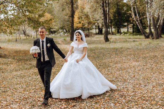 A wedding couple is walking in nature on an autumn day. Happy young bride and elegant groom holding hands. A stylish couple of newlyweds on their wedding day.