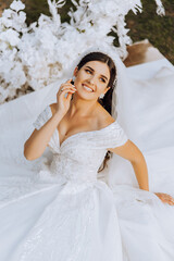 The brunette bride, smiling sincerely, sits on the ground in a unfolded dress, near a white arch...