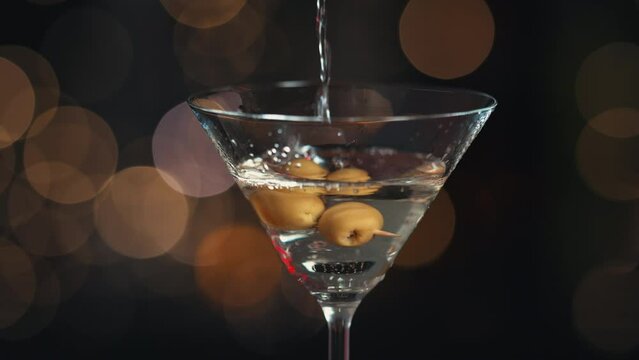 Llena la copa de Martini con alcohol líquido, tiene dos aceitunas en un palillo. Todo el fondo está desenfocado, creando un efecto bokeh con las luces de fondo. Primer plano del relleno de vidrio.