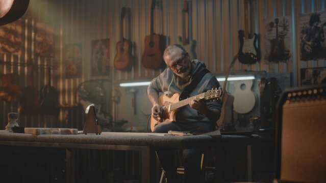 Mature musician plays guitar in stylish workshop for making musical instruments. Professional carpenter or craftsman checks the guitar after production. Handcrafted wooden guitars hang on the wall.