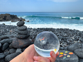 Hand holding crystal ball, zen pyramid and chakra healing stones on volcanic ocean shore.