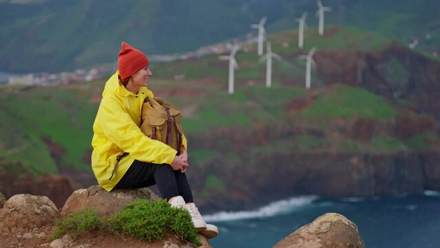 Attractive traveling woman in a yellow jacket and backpack looking at windmills in field 