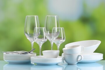 Set of many clean dishware, cutlery and glasses on light blue table against blurred green background