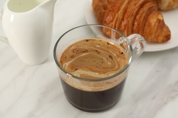 Aromatic coffee in glass cup, fresh croissant and pitcher on white marble table, closeup