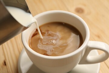 Pouring milk into cup with coffee at light wooden table, closeup