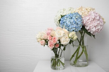 Beautiful hydrangea and rose flowers in vases on white bedside table indoors, space for text