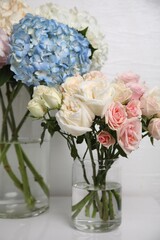 Beautiful hydrangea and rose flowers in vases on white table, closeup