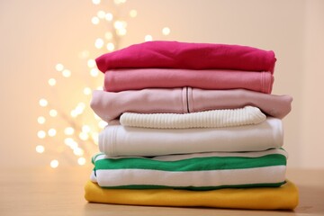 Stack of folded clothes on wooden table against beige background with blurred lights