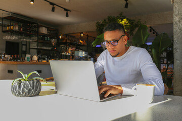 Young latin guy with laptop working remotely from a coffee shop.