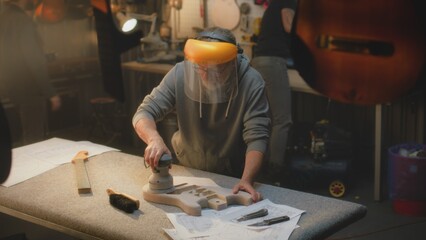 Caucasian mature artisan in protective mask grinds guitar body using grinding machine. Craftsman...