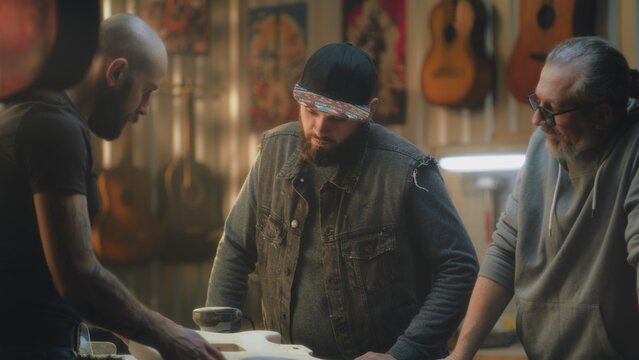 Male professional artisans discuss work and check blueprints in light stylish workshop. Tattooed carpenter makes electro guitar and talks with colleagues. Concept of handcraft and small business.