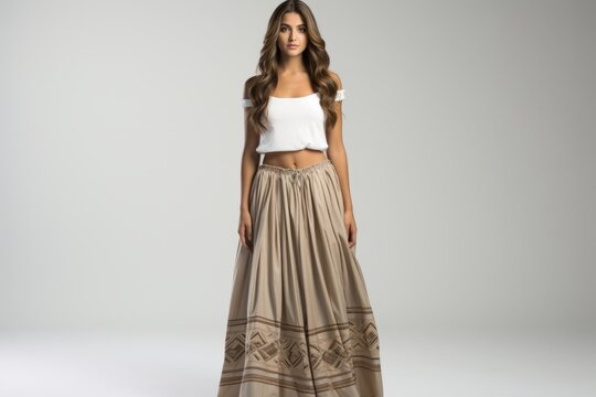 Fashion model with long brown hair wearing a white off-shoulder crop top and a beige pleated maxi skirt with brown geometric embroidery