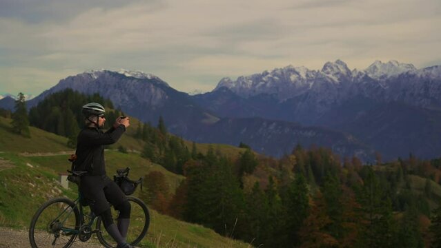 Male cyclist takes photos on smartphone in Bavarian Alps in Germany. Bicyclist shoots video of snow covered mountains. Male cyclist on gravel bike enjoying view and making phono on top of mountain. 