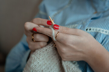 hands of woman with nail polish and rings knitting a beige scarf with knitting needles during hte...