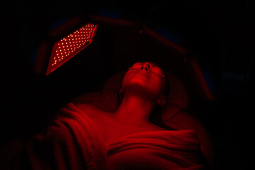 Top view of relaxed female client having red LED light facial photodynamic therapy treatment in...