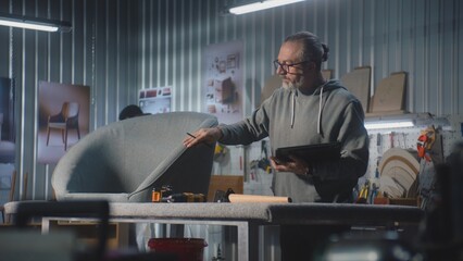 Mature carpenter or designer uses digital tablet and works with chair in modern workshop for making and renovation furniture. Another specialist assembles shelf. Handmade and entrepreneurship concept.