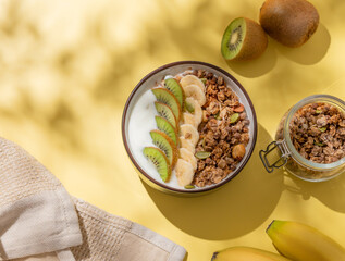 Homemade granola with banana, kiwi and nuts in a bowl on a yellow background with fresh fruits and shadows.