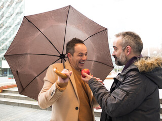 Two forty year old executives with umbrellas offer each other a red apple on a rainy winter street symbolizing mutual respect and camaraderie - 741848089