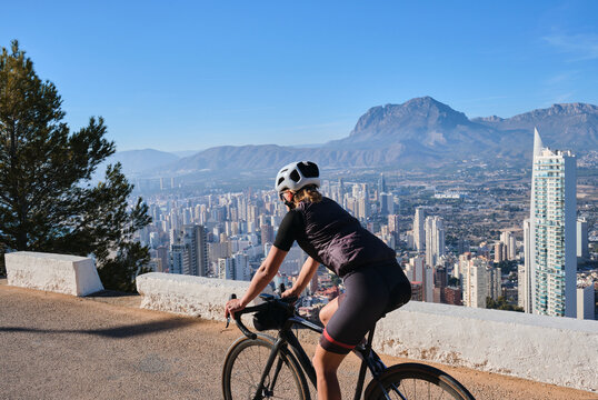 Woman cyclist riding a bike with beautiful view on Benidorm,Costa Blanca,Spain.Woman cyclist wearing cycling kit and helmet.Sports motivation image.Cycling through stunning Spanish mountain landscape.