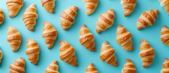 The image showcases numerous freshly baked croissants with a golden-brown crust evenly spaced out on a vibrant blue background. The pastries exhibit a flaky texture and appear to be made with a butter - Powered by Adobe