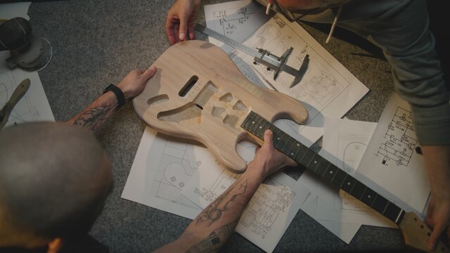 Top view of two male carpenters talking, making guitar according to drawings and taking measurements. Craftsmen work in modern workshop for musical instruments making. Handmade and entrepreneurship.