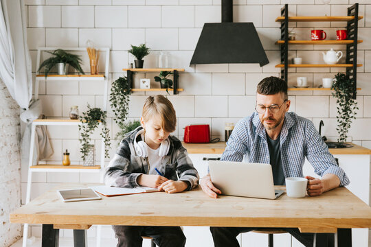 family dad young man with phone and son teenage cute boy with tablet and headphones doing homework sit at table in cosy apartment, quality time with your family, gadget addicted, education