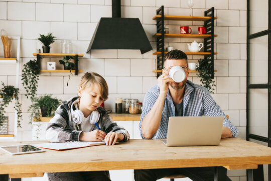 family dad young man with phone and son teenage cute boy with tablet and headphones doing homework sit at table in cosy apartment, quality time with your family, gadget addicted, education