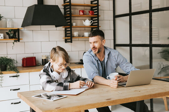 family dad young man with phone and son teenage cute boy with tablet and headphones doing homework sit at table in cosy apartment, quality time with your family, gadget addicted, education