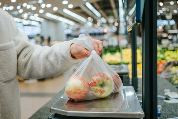 A person's hand is shown placing a plastic bag of fresh apples on a digital scale in the produce...