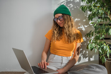 A stylish freelance woman with a laptop is sitting on the floor of a green ficus tree at home and...