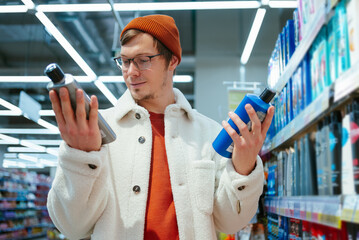 A discerning shopper in a fleece jacket and beanie compares two shampoo bottles in a store aisle,...