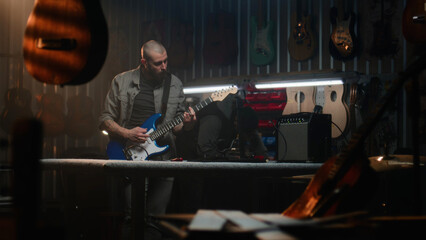 Male craftsman plays guitar connected to combo amp, checks the sound. Carpenter makes wooden...