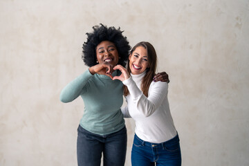 Studio shot of a two diverse young women forming a heart shape while standing against a background...