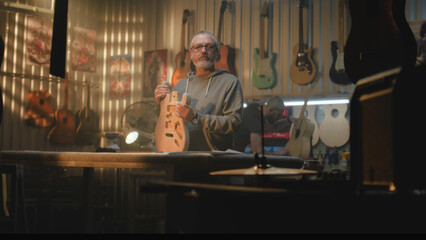 Fototapeta na wymiar Mature carpenter, craftsman stands in modern woodworking workshop, holds wooden guitar body in hands and looks at camera. Colleagues make musical instruments. Handmade and entrepreneurship. Portrait.