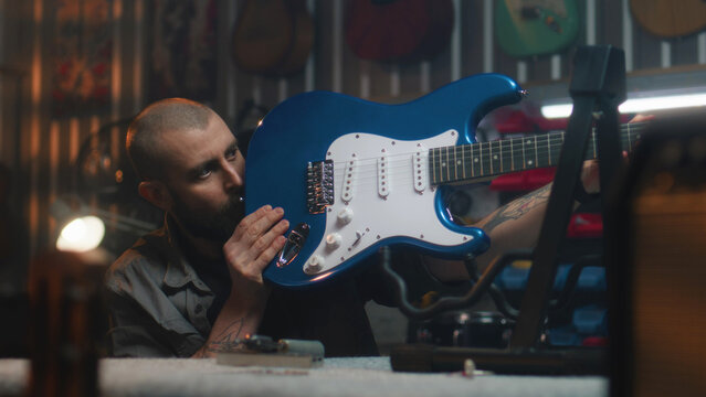 Male carpenter detailed inspects the guitar and strings. Craftsman makes musical instrument from wood in modern workshop. Another artisan works in the background. Handcraft and small business concept.
