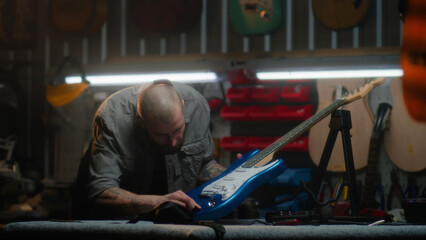 Male carpenter inspects guitar in detail and wipes with it rag. Craftsman makes wooden musical...