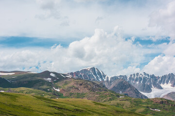 Scenic alpine landscape with green hills and rocks with view to large snow-capped mountain peak...
