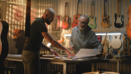 Fototapeta na wymiar Two professional carpenters, craftsmen inspect wooden guitar body blank, discuss process of making musical instrument in modern workshop. Colleague works in background. Handmade and entrepreneurship.