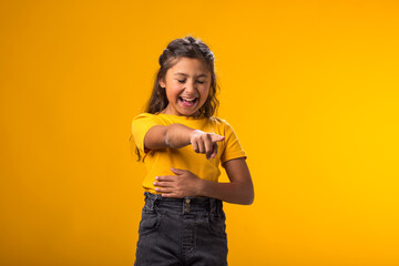 Kid girl mocking and teasing at someone showing finger at camera and holding stomach over yellow...