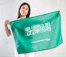 Cheerful young woman standing with big flag of Saudi Arabia against light shadeless background