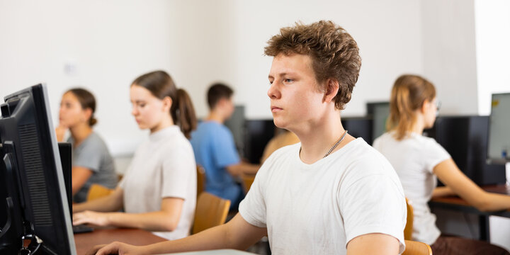 Focused teenager male student sitting at desk in college computer class, preparing for exam