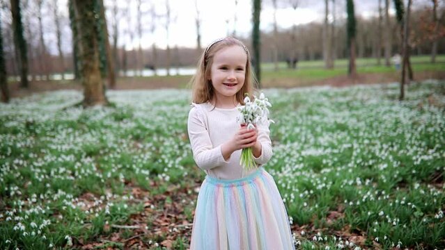 Cute preschooler girl in green tutu skirt gathering snowdrop flowers in park or forest on a spring day. Little kid exploring nature. Outdoor activities for children