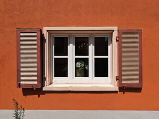 A view into a rustic window and shutters affixed to a stucco home. Orange in color with a grey strip at the bottom. A small plant grows. Looking through the window to a house plant and light fixture.